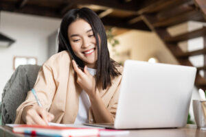 smiling-asian-woman-working-from-home-taking-notes-notebook-talking-cellphone-writing-down